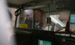 Taxi driver behind glass partition in New York City, focused on urban commute.