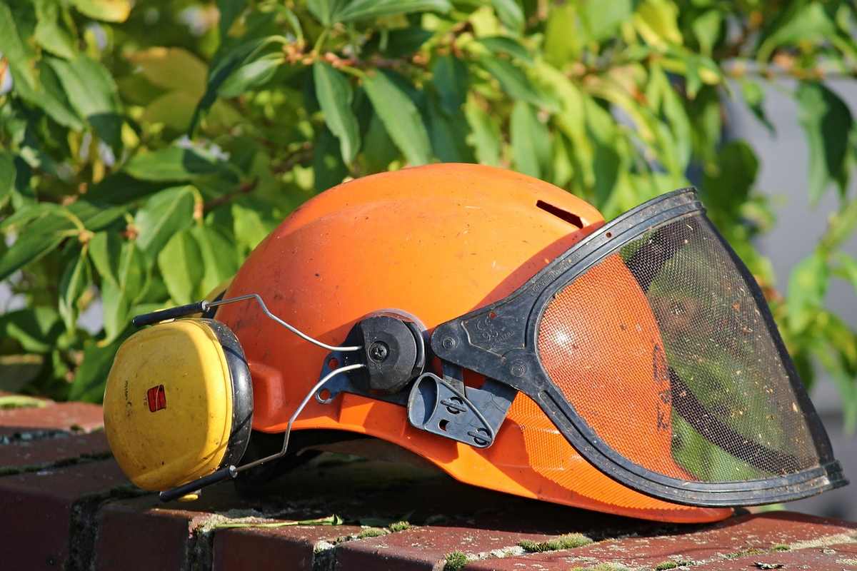 Casque de sécurité avec visière et protections auditives, posé sur un mur de briques, devant des feuilles vertes